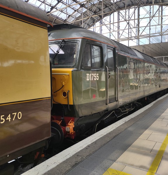 An old diesel train in its platform at Queen Street station. The train is blocky and painted olive green,  the end of an old train carriage is visible in front, painted brown and cream.  The platform is paved and level, with a row of tactile tiles, a white edge and painted yellow guidelines. The arching glass roof of the station and concourse area are visible above and beyond.