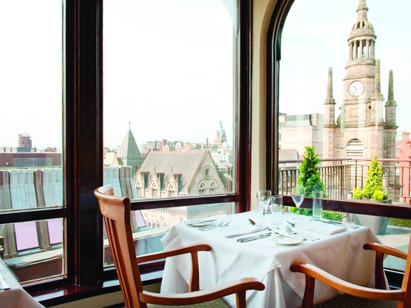 An internal view of the Carlton George shows tables and chairs arranged for dinner. Each wall is lined with windows. Outside the window, the roof-terrace railings are visible and beyond them a steeple and the rooves of old and modern buildings can be seen against a blue sky