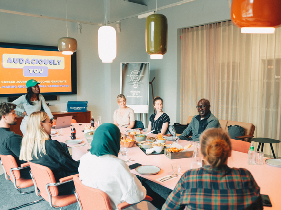 Group of people sitting around an oval boardroom table. Person standing at top of table in front of a large screen that reads 'Audaciously You'