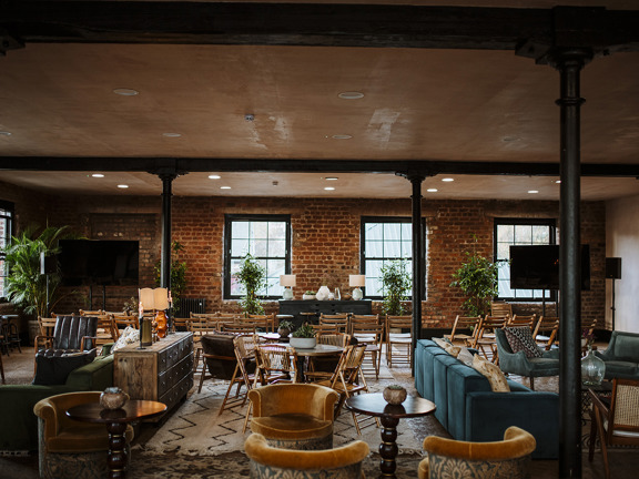 An interior image of The Engine Works, Print Room. The image shows an ex-industrial space with a mix of red brick and gypsum walls, the ceiling is supported by slim cast iron pillars painted black. The far and left-hand walls have large, paned windows with black frames.  The room is filled with an eclectic mix of furnishings, including rugs, velvet armchairs, plants, round wooden tables, table lamps and wooden folding chairs. Speakers and 2 large TV screens on stands flank the room on either side.