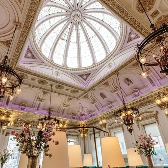 An interior view of Corinthian Club looking up at a large glass dome. Opulent, decorative plaster work is visible around the dome and across the entire ceiling in white and gold. The room is very bright with daylight streaming through the dome as well as the windows that cover both visible walls. There are also chandeliers and more modern light fittings visible. Large vases of foliage and pink flowers are also visible.