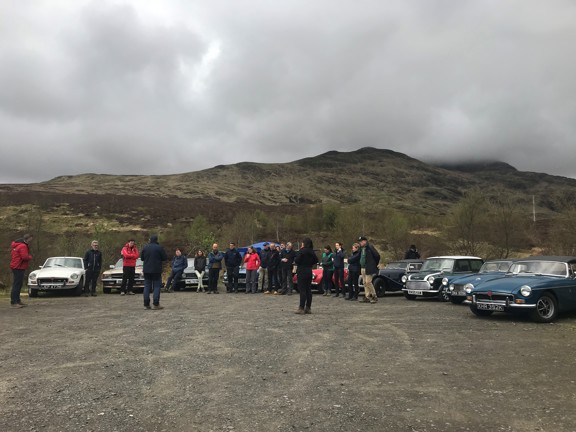 Group of people standing in front of vintage cars in the Scottish countryside with mountains surrounding the group. 