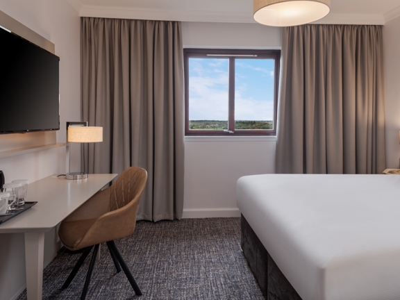 An interior view of a DoubleTree by Hilton Glasgow Westerwood bedroom. The room is light, with a small window and decorated with light coloured furnishings. A desk, tan leather chair, kettle and wall-mounted television are all visible.