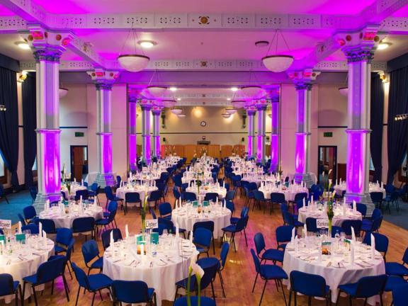 Room set up with round tables for dinner, white linen table coverings with blue chairs around the tables. Ornate pillars in the room are washed in pink/purple light.