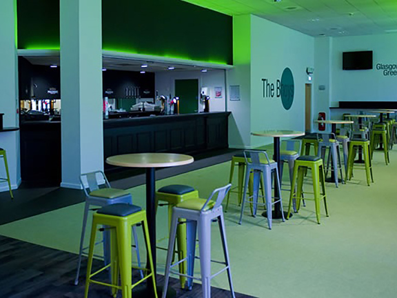 An internal view from Celtic Football Club shows a room dotted with high, round tables and metal stools in shades of green and grey. A dark bar takes up the length of the far wall, the bar and around the ceiling is lit with green neon. Wall decals read "The Bhoys" and "Glasgow's Green and White".
