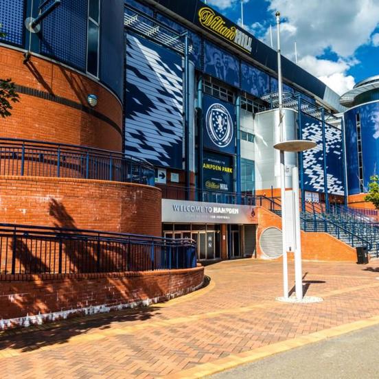 An exterior view of Hampden Stadiums main entrance. A large brick and blue metal clad building, with round towers at the two visible corners. The facade is decorated with sponsors, the Scottish FC badge and lettering in white or yellow reading," Hampden Park."  The area outside of the main entrance is paved with brick and curves down to 3 or more doors. Adjacent steps lead up to a further entrance, furthermore, ramps curve out of sight around the towers' bases. Trees and modern streetlights line the path.