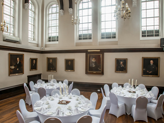 An interior view of a room at 1599 filled with round tables surrounded by chairs. Candles and napkins decorate the tables. The room has wooden floors, arched windows and a vaulted ceiling.