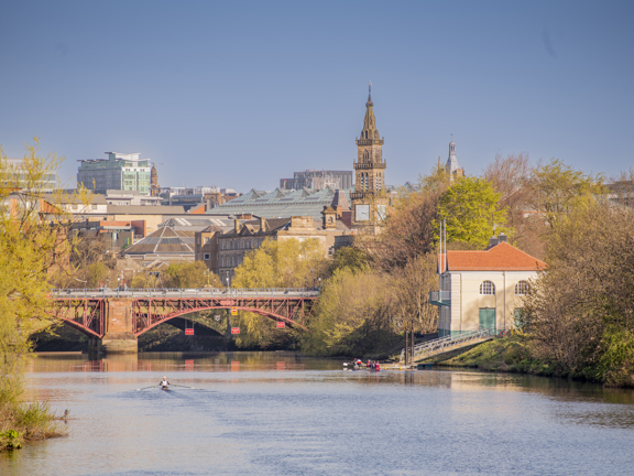 Sunny view of rowers on single sculls on the tree-lined River Clyde with a bridge and the city in the background