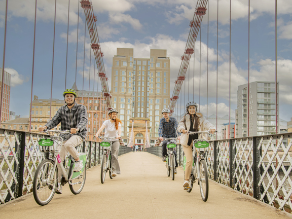 Four cyclists crossing a footbridge with the modern grey building of the Virgin hotel in the background