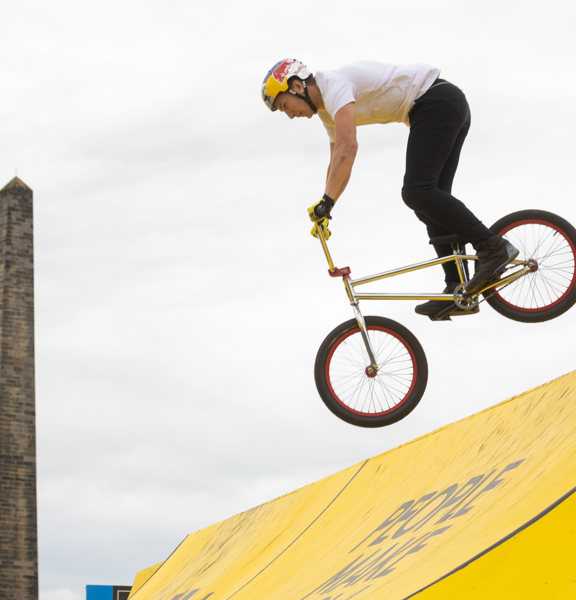 BMX Cyclist rides down yellow slope, with Nelson Monument, Glasgow in the background.