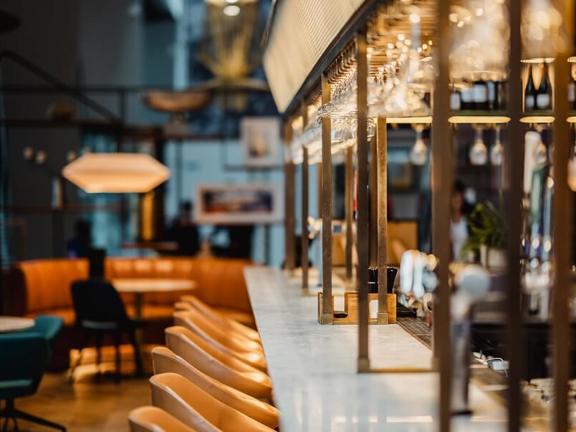 Marble topped bar with brass metal framework above and beige leather barstools lined up along one edge.