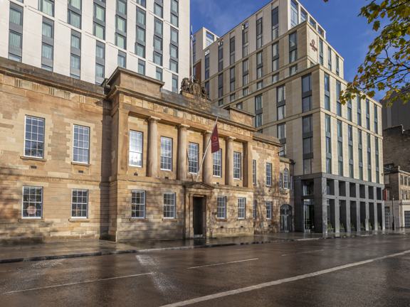 An exterior view of the Clayton Hotel shows a golden-sandstone building with carved details and 4 Greek Doric style pillars adorning the front. A few steps lead up to the main entrance above which a maroon flag with the orange Clayton logo hangs. Behind the sandstone building a modern, cream and grey, multi-storey building is visible against blue skies.