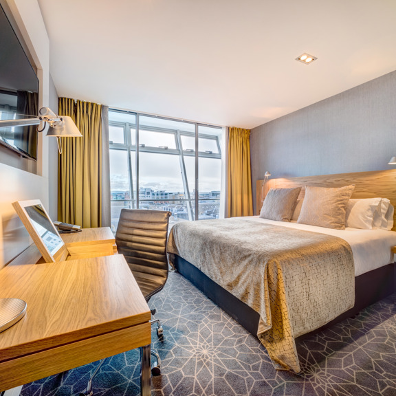 An interior shot of a hotel room, the bed is large and tidy and has a grey blanket and cushion on it. A television, desk and chair are all visible. The far wall is floor to ceiling windows with yellow curtains on either side.