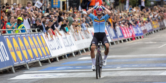 Woman cyclist crosses race finish line with hands on head, in front of cheering crowds in Glasgow.