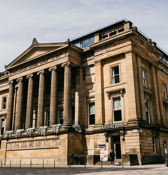 An exterior view of Citation shows an imposing, golden-sandstone building adorned with carved details and 6 large, Greek Ionic style pillars. Windows are visible on 4 storeys and grey lettering reading "Citation" is visible above doors on either side of the pillars.