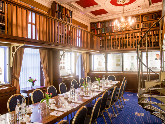 A room at 1599 set out with a long table in the middle, pens, paper and bottled water are visible on the table. Around the edge of the room display cases and book cases are visible. A spiral staircase up to a mezzanine balcony is also visible.