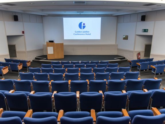 An interior image of a Golden Jubilee hotel lecture theatre. The image is taken from the back of the room from the top of the tiered theatre-style seating. The seats are blue with wooden arms and swivel desks. At the front of the room a wooden lectern with a screen and microphone is positioned in front of a projected image of the hotel's branding. Speakers can be seen on either side of the room above doorways out of the room. 