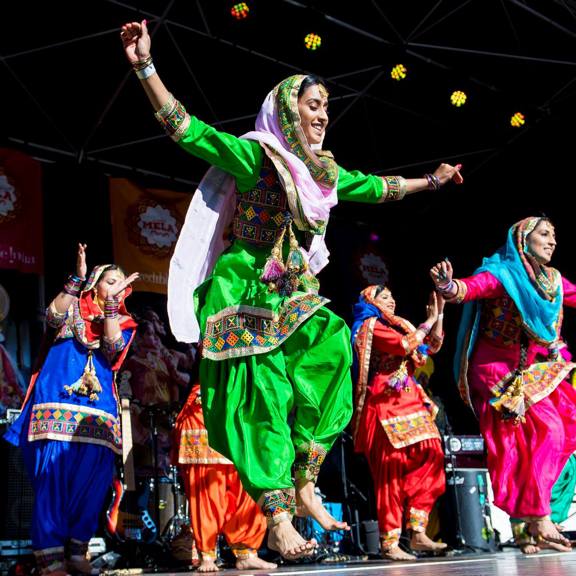 A group of women dancers in traditional clothing dance on stage in the sunshine at Glasgow Mela.