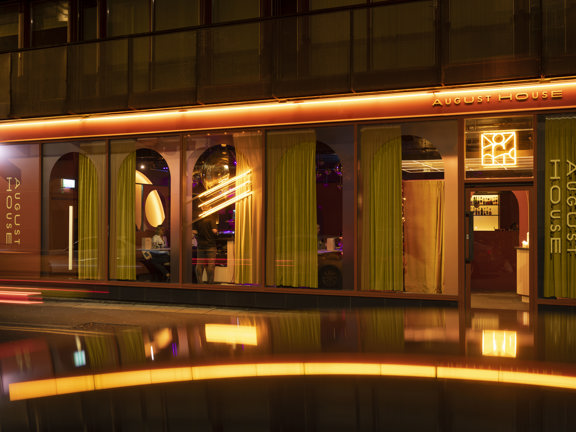 An exterior shot of August House shows a wide ground floor business with plate glass windows. Stylised, yellow, vertical vinyl lettering on the building and windows read "August House". It is evening and lights from with the bar make heavy yellow velvet curtains and a disco ball visible within.