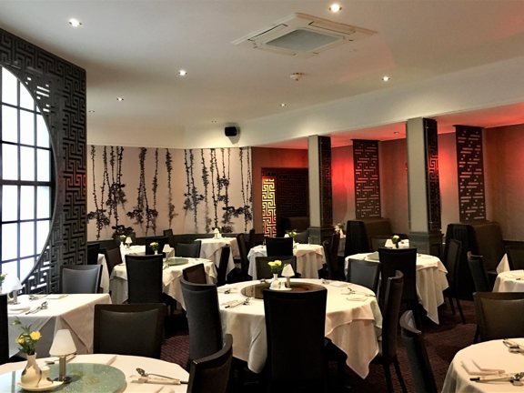 An interior view of the main dining room at Amber Regent. A large dining room with patterned, dark-red carpet and mainly cream walls. A monochrome botanical wallpapered wall and lattice work screens add dramtic accents to the room. The left wall also has a large, paned circular window. A dozen round tables, covered in white cloths and laid for dinner are scattered around the room, surrounded by dark upholstered, high-backed chairs.