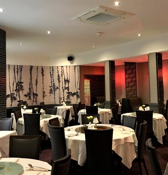 An interior view of the main dining room at Amber Regent. A large dining room with patterned, dark-red carpet and mainly cream walls. A monochrome botanical wallpapered wall and lattice work screens add dramtic accents to the room. The left wall also has a large, paned circular window. A dozen round tables, covered in white cloths and laid for dinner are scattered around the room, surrounded by dark upholstered, high-backed chairs.