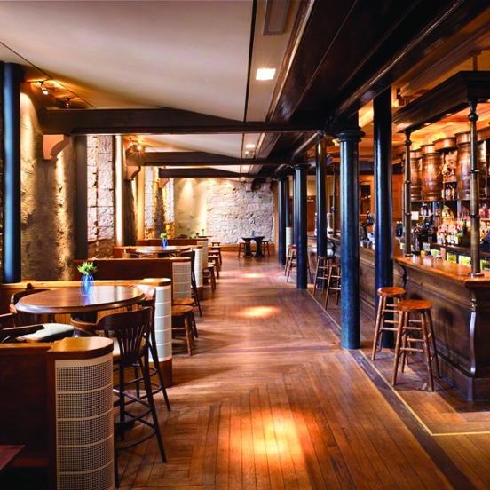 An interior of Oran Mor's bar space shows a room with bare stone walls and a low ceiling with wooden beams and black steel columns running the length of an old fashioned, wooden, panelled bar. The bar stretches the full length of the space, the fridges and shelves behind are fully stocked with bottles and 4 old fashioned wooden barrels are also visible on the top shelf. Tall wooden stools are placed intermittently along the bar, on the left tables with stools and chairs are placed beside each window.