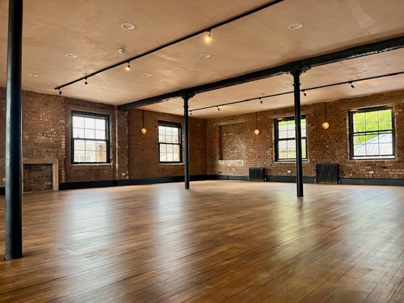 Post industryial room with wooden floor boards, Steel columns supporting the room, four black framed windows on the far side of the room allow views to the garden beyond. 