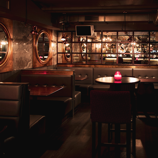 An interior from Blue Dog of booth seating, with high-backed leather upholstery around dark wooden tables. 2 large round mirrors with copper surrounds adorn the wall above each visible booth. The far wall is mirrored showing a reflection of a lit bar. Speakers and lights are mounted to the walls. 