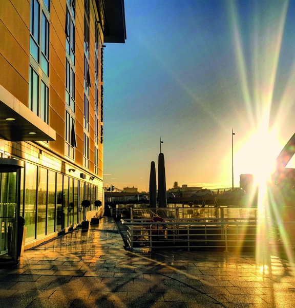 An early morning shot of the Hilton Garden Inn terrace shows the exterior of the hotel and the Clyde Arc bridge lit by the sun. The sun is low in the sky and its rays penetrate the image. The hotel ground floor is windowed floor to ceiling along the length of the visible wall. A glass revolving door with red lettering above, reading "Hilton Garden Inn" can be seen in the foreground. The paved floor is wet from rain and reflects the suns light. White railing run along the side of the River Clyde.