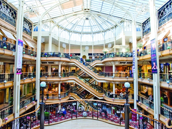 An interior image of the vast, tiered interior of Princes Square and its impressive pitched glass roof. Curving wooden balconies with black railing and sleek elevators, sweep around the outside edges of the open atrium. Awning, signs and banners advertise the shops, restaurants and businesses housed within Princes Square. It is a bright day and daylight spills through the roof. 