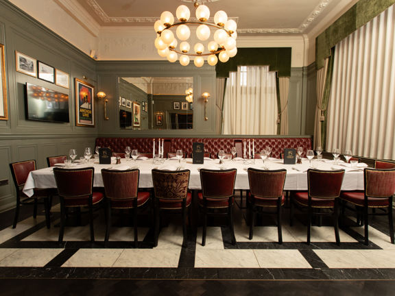 Interior of a restaurant private dining room with one long table across the middle of the room. The table is flanked by dark red chairs and leather banquettes. 