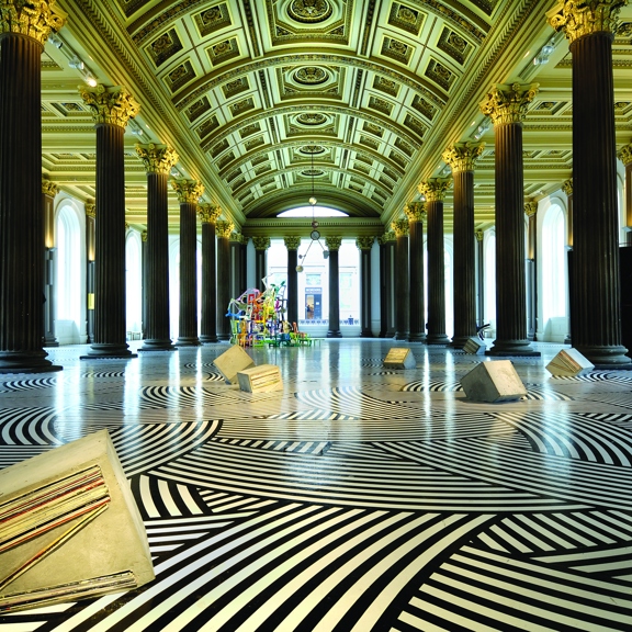 An interior shot of The Gallery of Modern Arts, Gallery 1. The image shows a large room lined with large arched windows. 2 rows of dark Roman-style columns line the room, reaching up to the gold, arched ceiling covered in decorative plaster work. The art installation covers the floor in a striped black and white pattern and concrete shapes and a colourful, wiry sculpture can be seen in the distance. Spotlights can be seen between the columns and the outer walls are uplit.