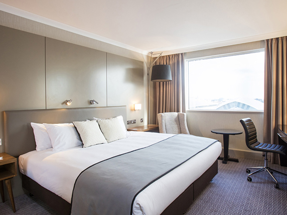 An interior view of a Crowne Plaza bedroom shows a hotel room decorated in neutral tones. The bed is large has clean white sheets at the foot of the bed there is a grey throw. A wooden bedside table, cream coloured armchair, black office chair and black desk are all partially visible. Plug sockets. a telephone, lamps and a television are also evident The beige curtains are pulled back but it is too bright to see the view out the window.