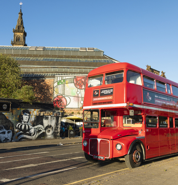 An image of the Red Bus Bistro, vintage double-decker bus parked beside Glasgow's famous Clutha Bar, its tree and its painted murals. It is a beautiful sunny day with blue skies. An old, stone spire and the grand, windowed roofline of the Briggait, a former fish market, re visible beyond. 