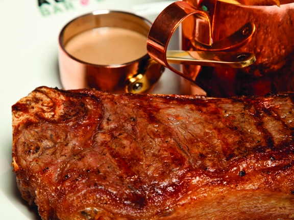 A close up image of an Alston Bar and Beef steak, it sits on a clean white plate. The rim of the plate is decorated with the Alston Bar & Beef logo of black text. Small copper containers hold golden French fries and a sauce,