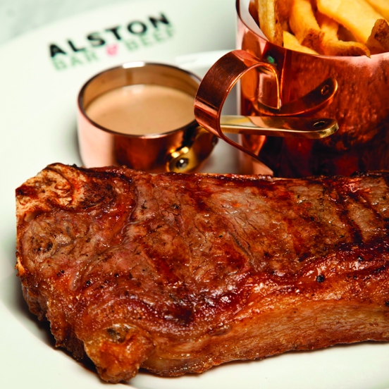 A close up image of an Alston Bar and Beef steak, it sits on a clean white plate. The rim of the plate is decorated with the Alston Bar & Beef logo of black text. Small copper containers hold golden French fries and a sauce,