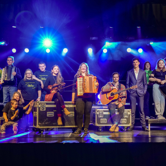 A group of musicians including men, women and children of different ages pose on a stage with musical instruments