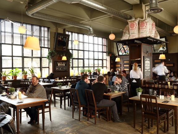 An interior view of the West on the Green dining room. A bright, open space with a post-industrial aesthetic. Large, paned, landscape windows fill much of the far, outside wall. The walls have dark panelling and the ceiling and top part of the visible walls is painted an olive green. The floor is made up of reclaimed wooden panels, with remnants of paint. Silver extraction tubes run across the ceiling. Wooden tables of different sizes are surrounded by traditional wooden dining chairs, occupied by diners.