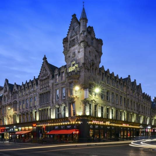 An exterior view of the Fraser Suites shows a Victorian sandstone building on a street corner, with a grand baronial inspired turret and carved details on the facade. The image is taken at dusk with a long exposure so streaks of light fill the road; streetlights and lights and signs from the ground floor businesses also light the scene. More modern buildings neighbour the Fraser Suite building at either end. 