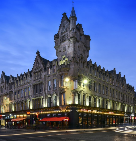 An exterior view of the Fraser Suites shows a Victorian sandstone building on a street corner, with a grand baronial inspired turret and carved details on the facade. The image is taken at dusk with a long exposure so streaks of light fill the road; streetlights and lights and signs from the ground floor businesses also light the scene. More modern buildings neighbour the Fraser Suite building at either end. 