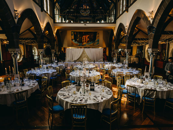 An interior image from Oran Mor taken from the back of an event space, decorated for a large, private dinner. 9 round tables, covered with white table cloths and set for dinner; with candelabras and silver wine buckets, fill the highly polished wooden floor of the main space. The side aisles are separated from the main nave by a series of pointed arches with stone pillars; the outer walls have circular stained-glass windows. A mezzanine level balcony is apparent, at the end of the room, above the top table.