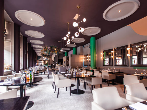 An image of the modern, bright dining room at Glaschu. A long large room with a dark, painted ceiling and cream carpets; plasterwork details on the ceiling and around the large windows in the left wall are painted white. 3 large, cylindrical pillars against the right wall are painted a bright, grassy green. The room is furnished with cream, leather chairs and glassy black tables. Modern spherical light features and a large dried-flower arrangement are suspended from the long ceiling at intervals.