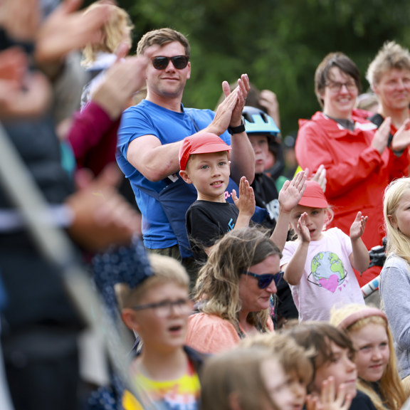 Several children and adults stand behind a barrier with T-shirts, hats and sunglasses on clapping for cyclists who are not in shot at the UCI Cycling World Championships 