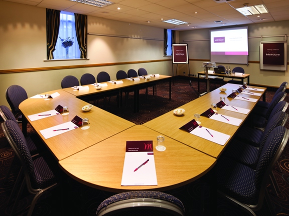 A Mercure hotel interior of the Broker Suite meeting room. The room is carpeted with a patterned brown carpet. The walls are cream, the ceiling is tiled and has fluorescent lights set into it. Tables with wooden tops are arranged into a U-shape, with fabric and metal chairs around the outside and pads and pens on top. At the far end of the room, a whiteboard, flipchart, a projector on a stand and a table with 2 chairs are all visible. To their left is 2 curtained windows with a vase of flower on each sill.