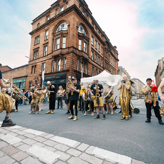 Around fifteen members of a brass band dressed in gold costumes play various instruments in the middle of the street during Merchant City Festival as an audience looks on