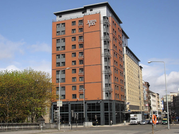 An external view of the Leonardo Hotel, shows a multi-storey, modern, red-brick building positioned on the corner of a junction. Metallic balconies punctuate the facade. The ground floor is windowed with dark glass and frames from floor to ceiling. A mixture of modern and Victorian buildings can be seen neighbouring the hotel on either side. A large, leafy tree and the carved stone of a bridge can be seen on the left. 