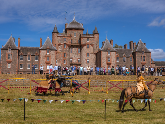 Large stone turreted building is the backdrop for jousting. A group of people line up to watch the joust while two people riding a horse charge at each other. 