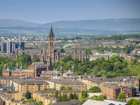 Sunny cityscape with the Gothic revival-style spire of the University of Glasgow's main building, surrounded by greenery and buildings of the West End, including the Baroque-style Kelvingrove Art Gallery and Museum