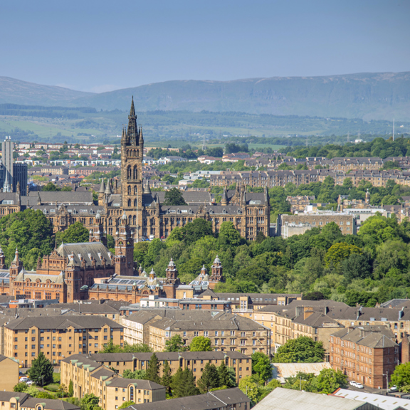 Sunny cityscape with the Gothic revival-style spire of the University of Glasgow's main building, surrounded by greenery and buildings of the West End, including the Baroque-style Kelvingrove Art Gallery and Museum