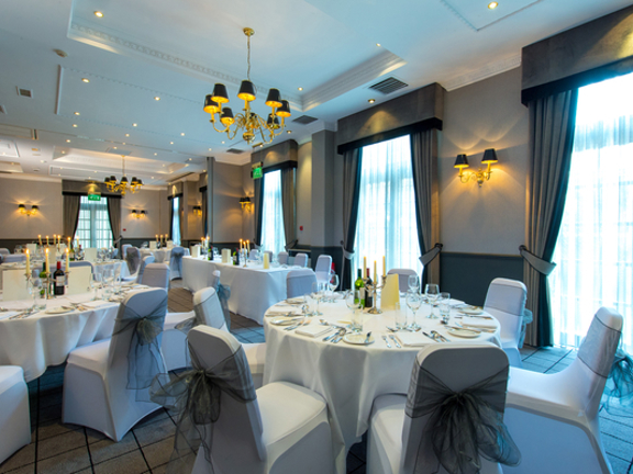 An interior shot of an event set up at Holiday Inn Theatreland shows a room with taupe walls and dark panelling, the floor is carpeted with a gridded pattern on it. The room is filled with round tables with white table cloths; wine bottle, candle sticks and glasses are all visible. White-covered chairs surround the tables, they are decorated with organza bows. Large French windows can be seen on the two visible walls, they are covered in white net curtains, dark grey curtains and pelmets frame them. 